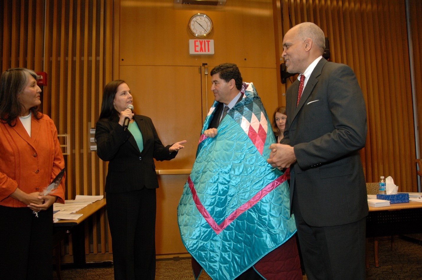 Dr Zerhouni is standing in a conference room wrapped in a star quilt, as a woman with dark hair holds a microphone, and appears to be singing