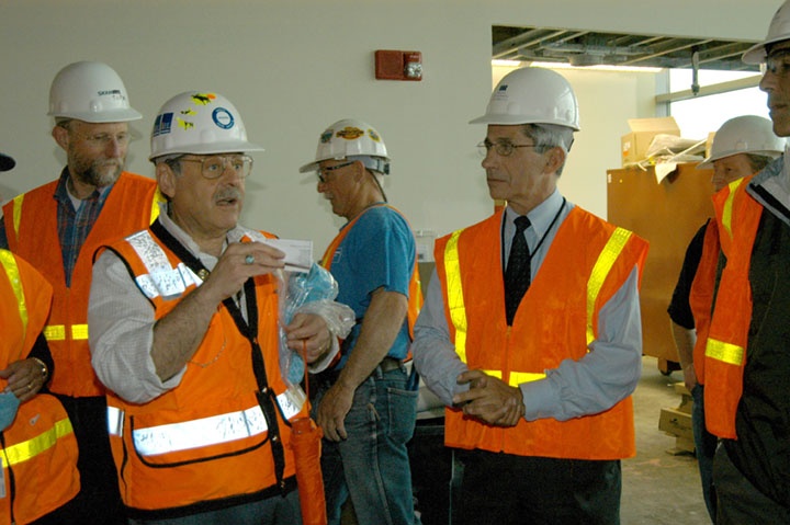 Marshall Bloom with Anthony Fauci during IRF Construction, June 2006. Both men, as well as those in the background, wear safety vests and hard hats. Bloom gestures to explain something to Fauci.