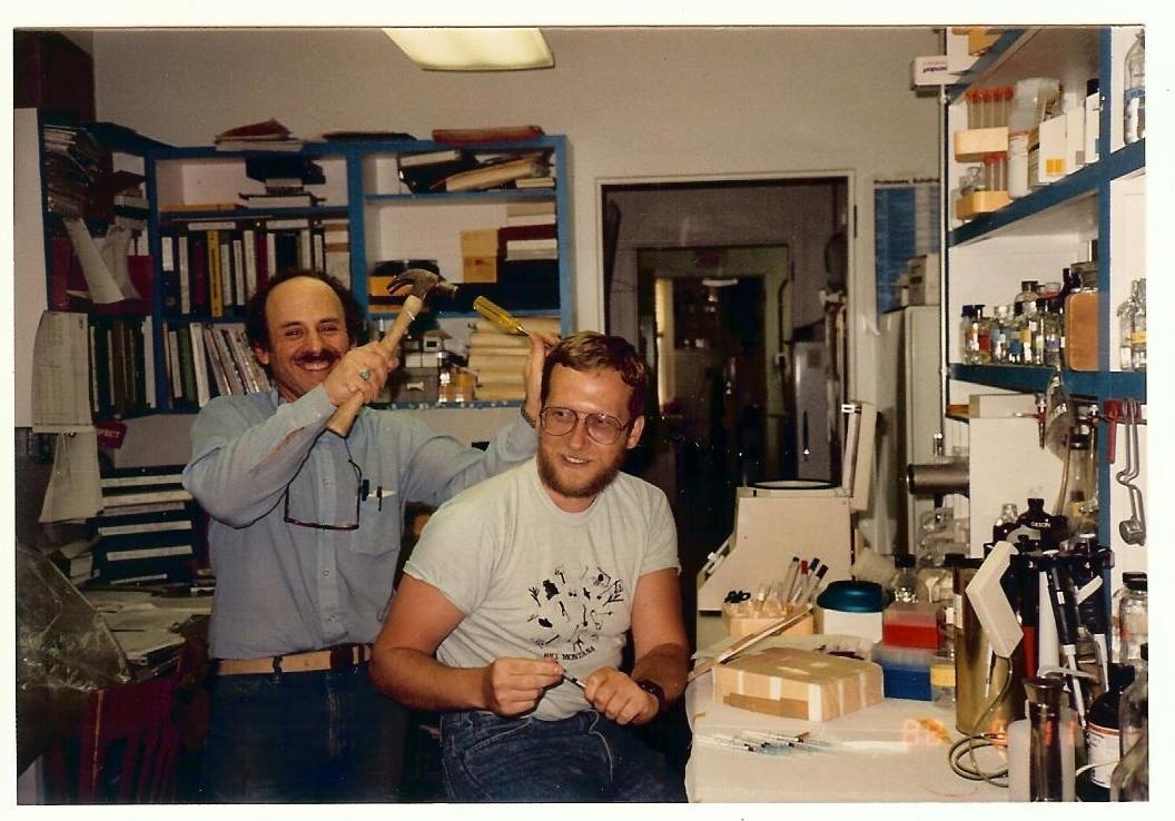 Marshall Bloom and Soren Alexandersen in the lab,  c. 1987. A smiling Bloom holds a chisel and hammer poised by Alexandersen's head, who is also smiling.
