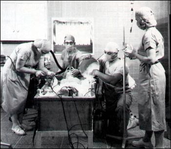 A photograph of open-heart surgery performed at the NIH Clinical Center using hypothermia. The patient was placed in a bed of ice to lower the total body temperature so that body tissues used very little oxygen. This permitted interruption of the blood flow for a brief period so that some procedures could be performed. This technique preceded the advent of the heart-lung machine, which today takes over the job of pumping blood during heart surgery.