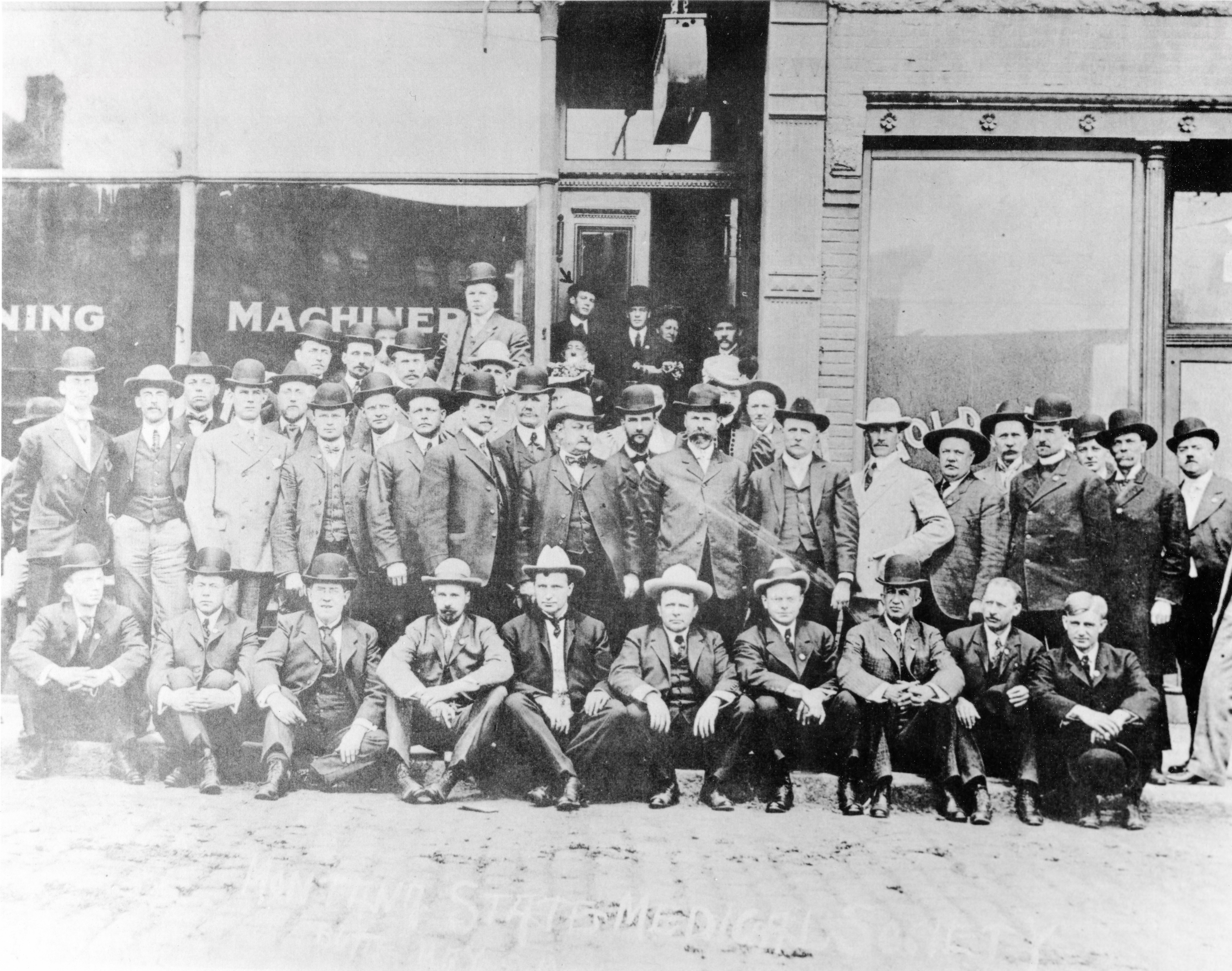 Several rows of men in bowlers pose on cobblestone street in front of a machinery shop. Rickets stands in the doorway at the back.