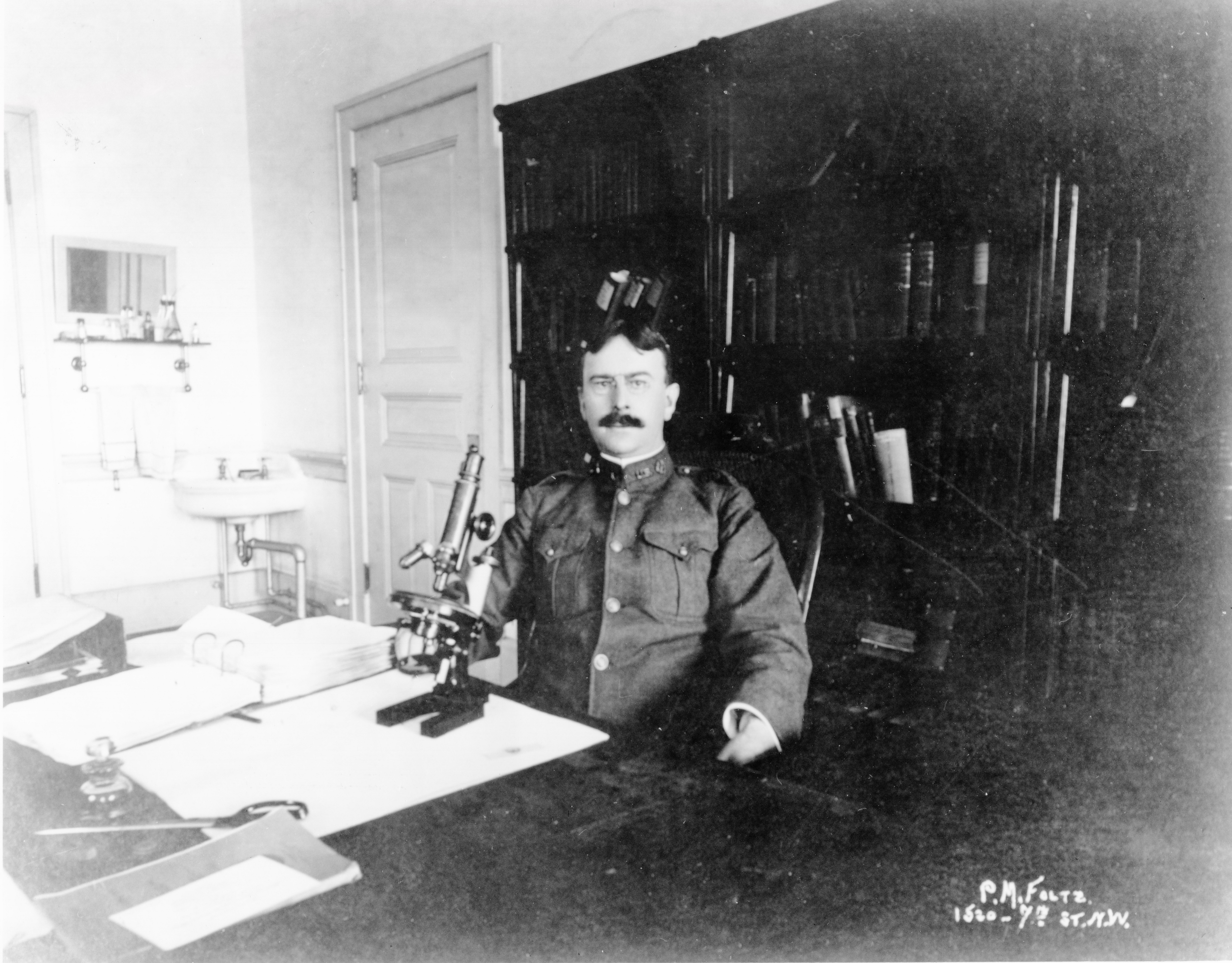 A man wearing a PHS uniform with a large mustache sits at a desk with a monocular microscope with a bookcase and sink in back
