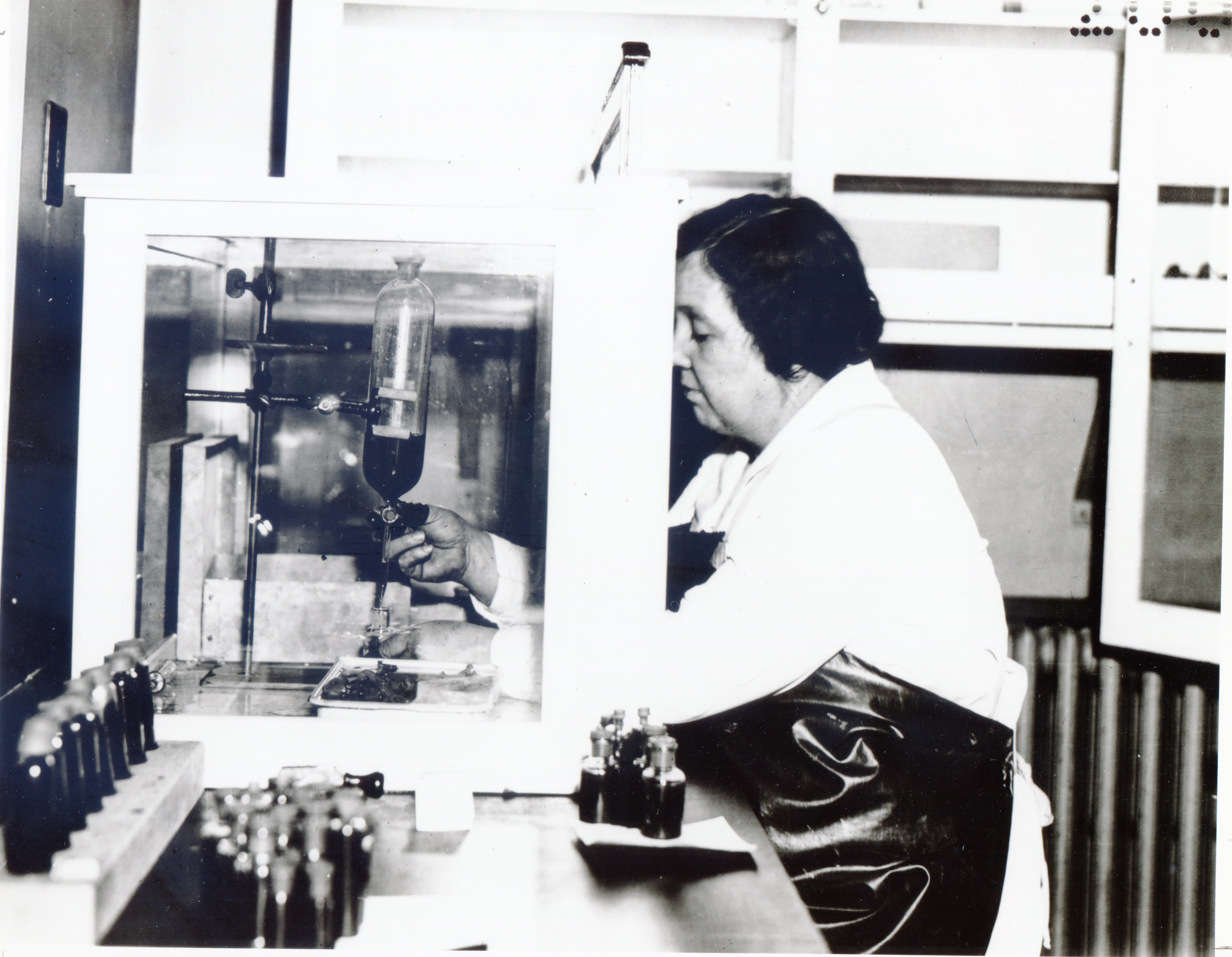 A woman bottles vaccine under a glass enclosure. She wears an apron but no protective gloves or mask.