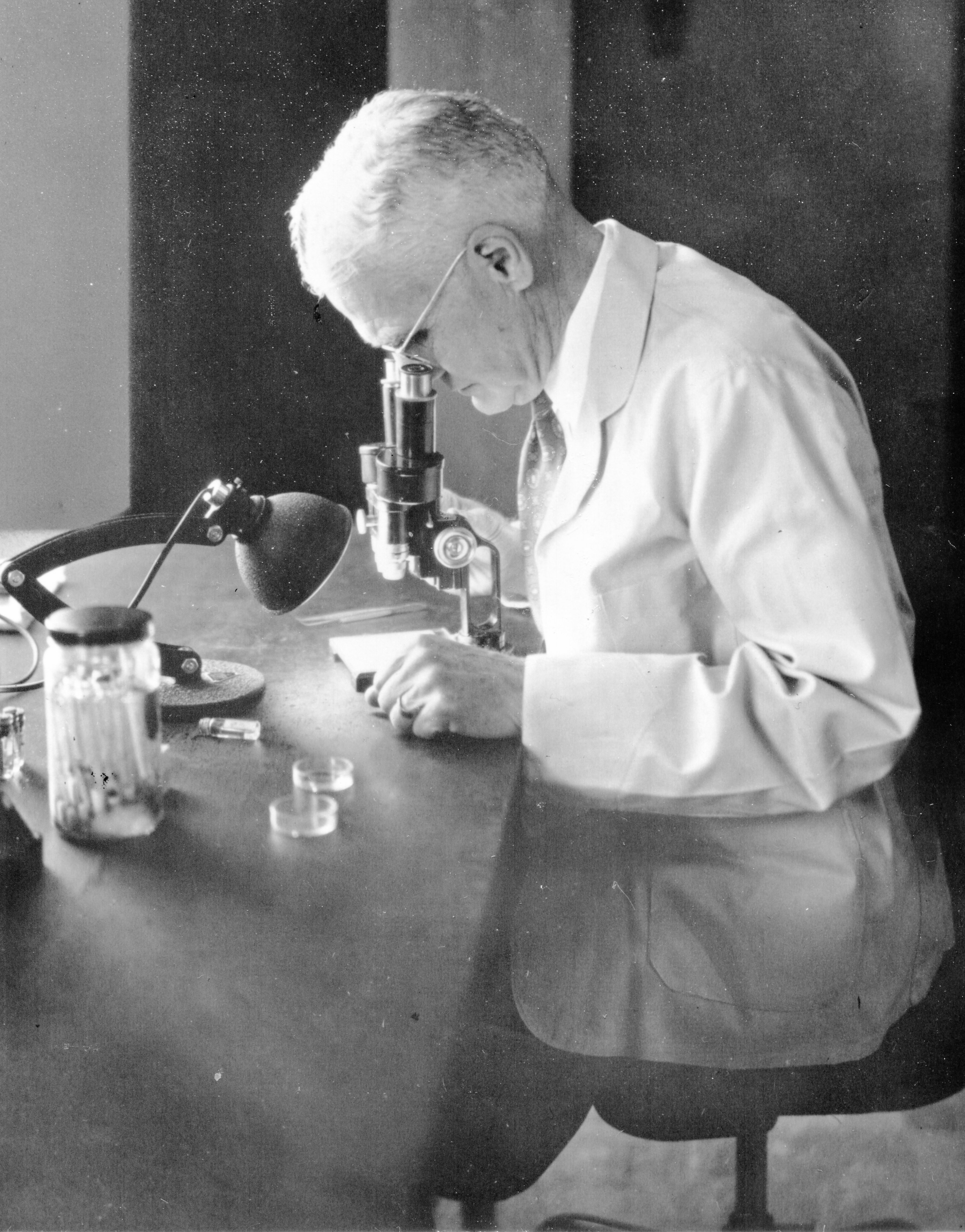 A white-haired Robert Cooley at microscope, in white coat, holding vials of ticks