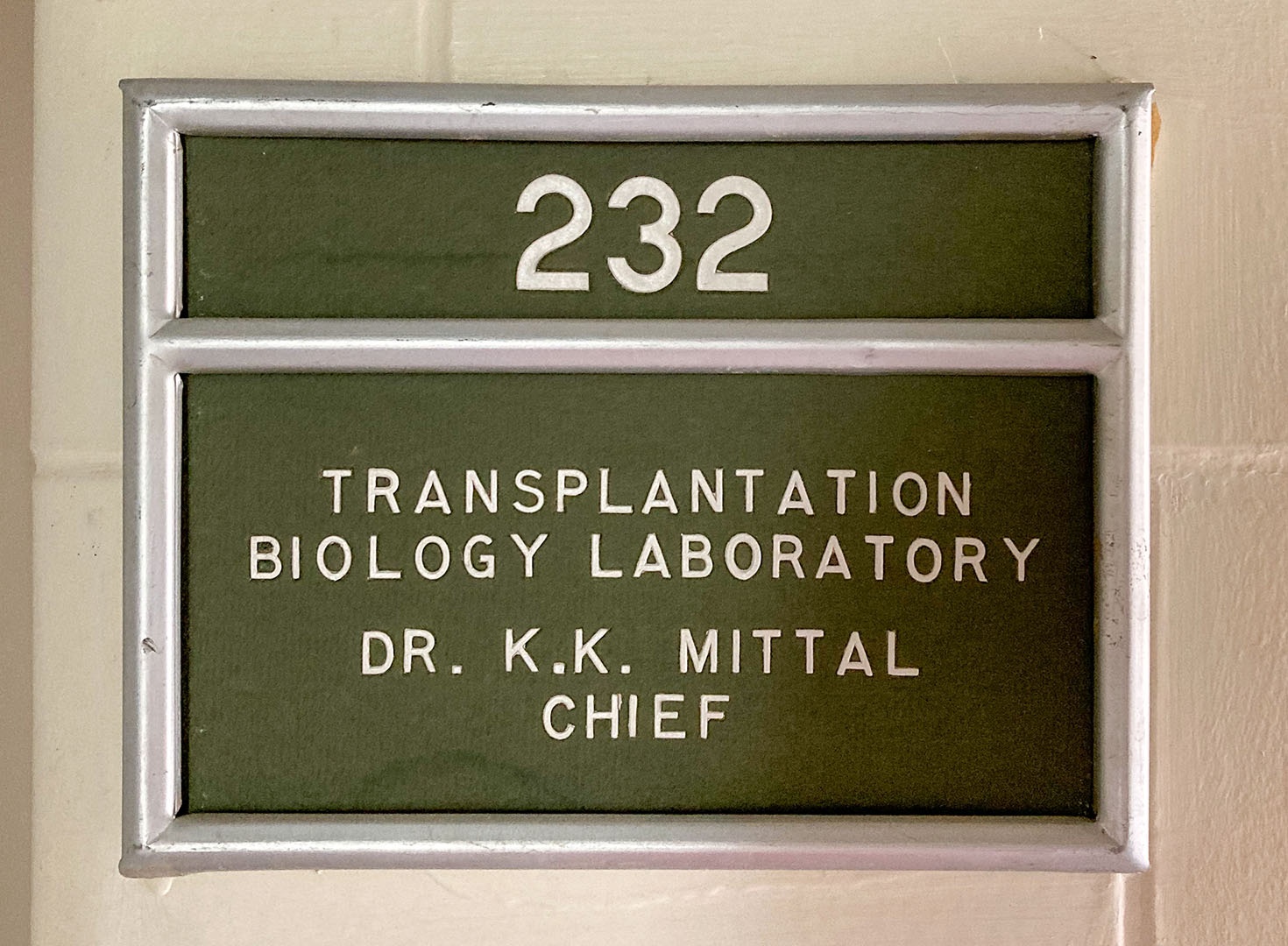A green and white sign with room number 232 and transplantation biology laboratory on it, in an aluminum rectangular holder