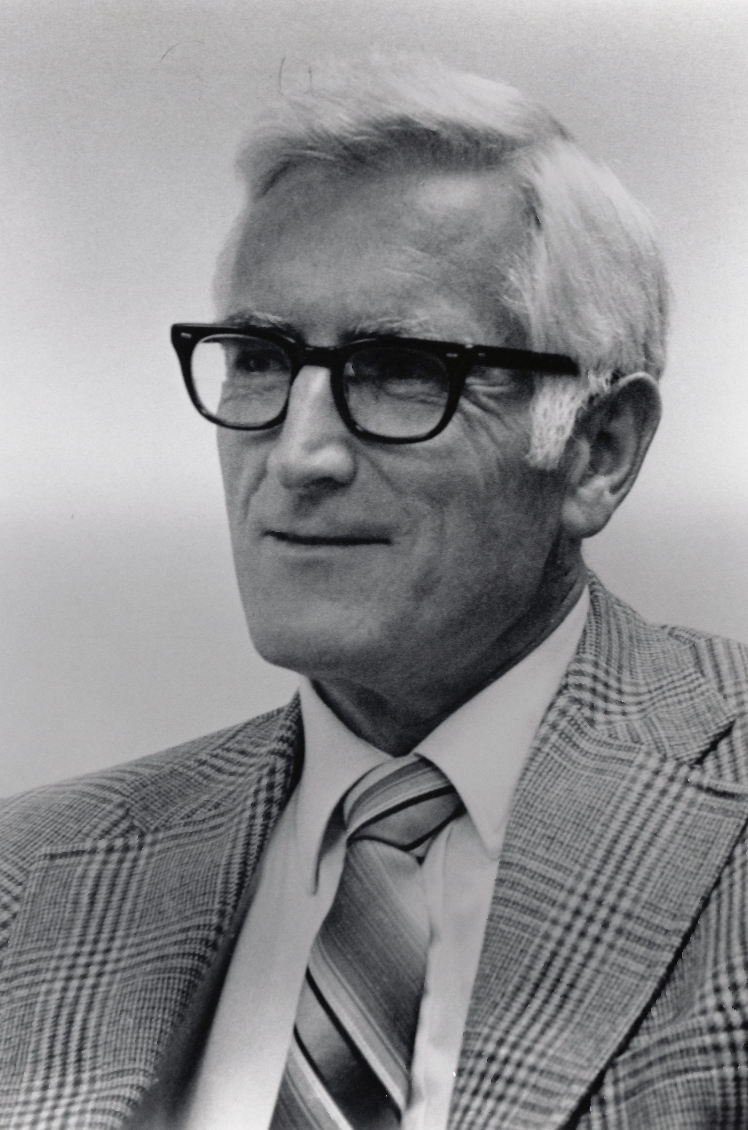a black and white photo of a man with black glasses wearing a plaid suit and tie