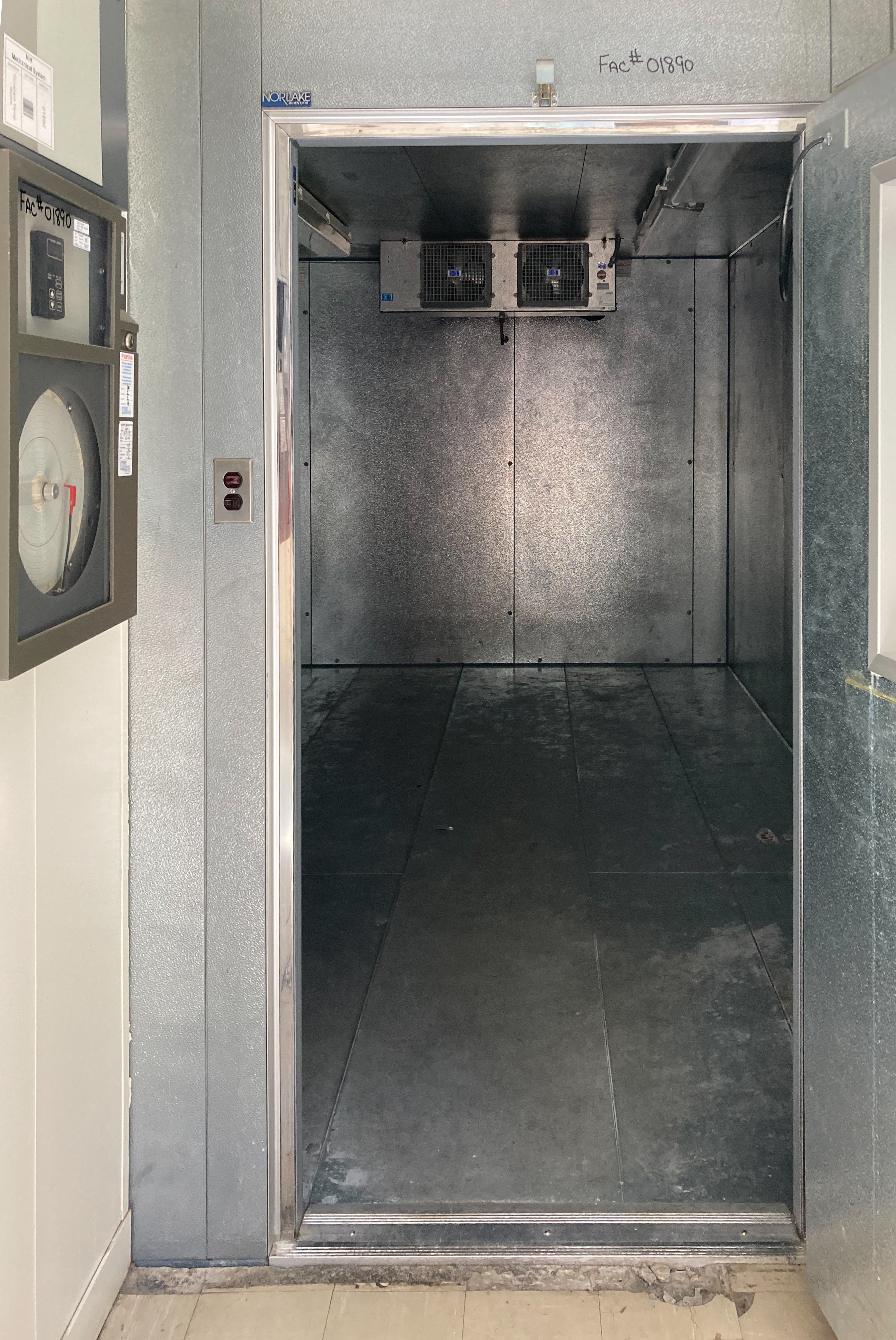 The refrigerated room with the door open. two fans can be seen in the top rear of the room, The walls and floor are metal. The room is empty.
