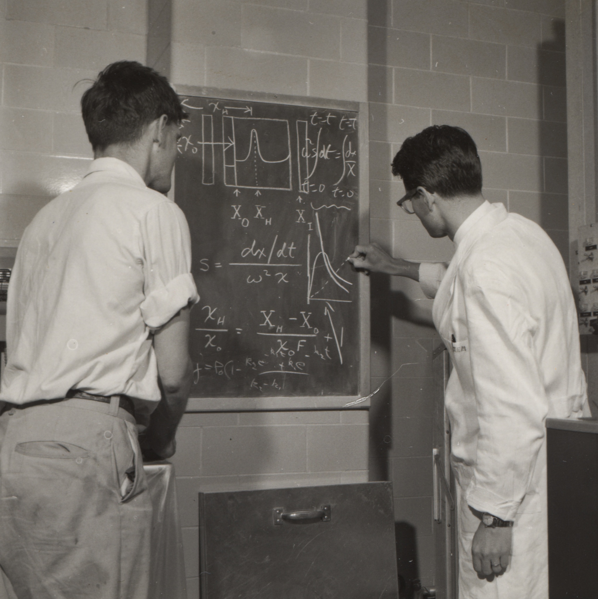 a black and white photo of two men working on an equation on a chalkboard. One man wears a white lab coat