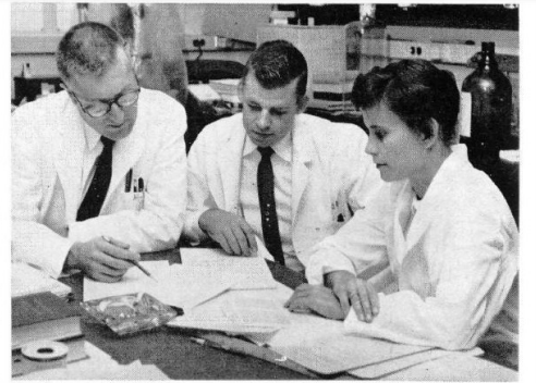 Two men and one woman seated at a desk in a laboratory, wearing white lab coats and looking at papers on the desk.