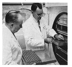 two doctors in lab coats examining a tray of tubes, half-inserted into a large device which resembles an oven.