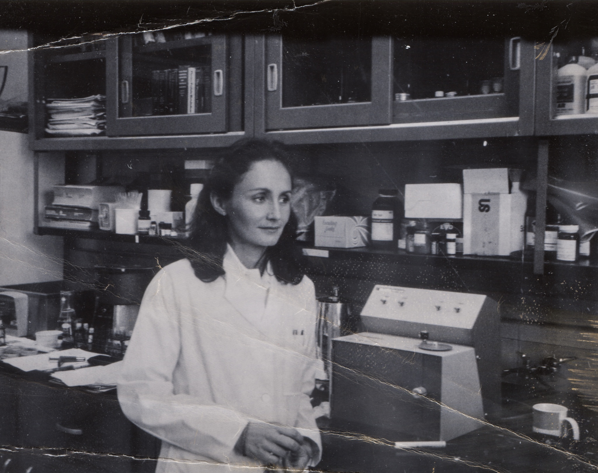 a black and white photo of a woman in a white lab coat next to a flurometer and coffee pot in a lab in 1974 