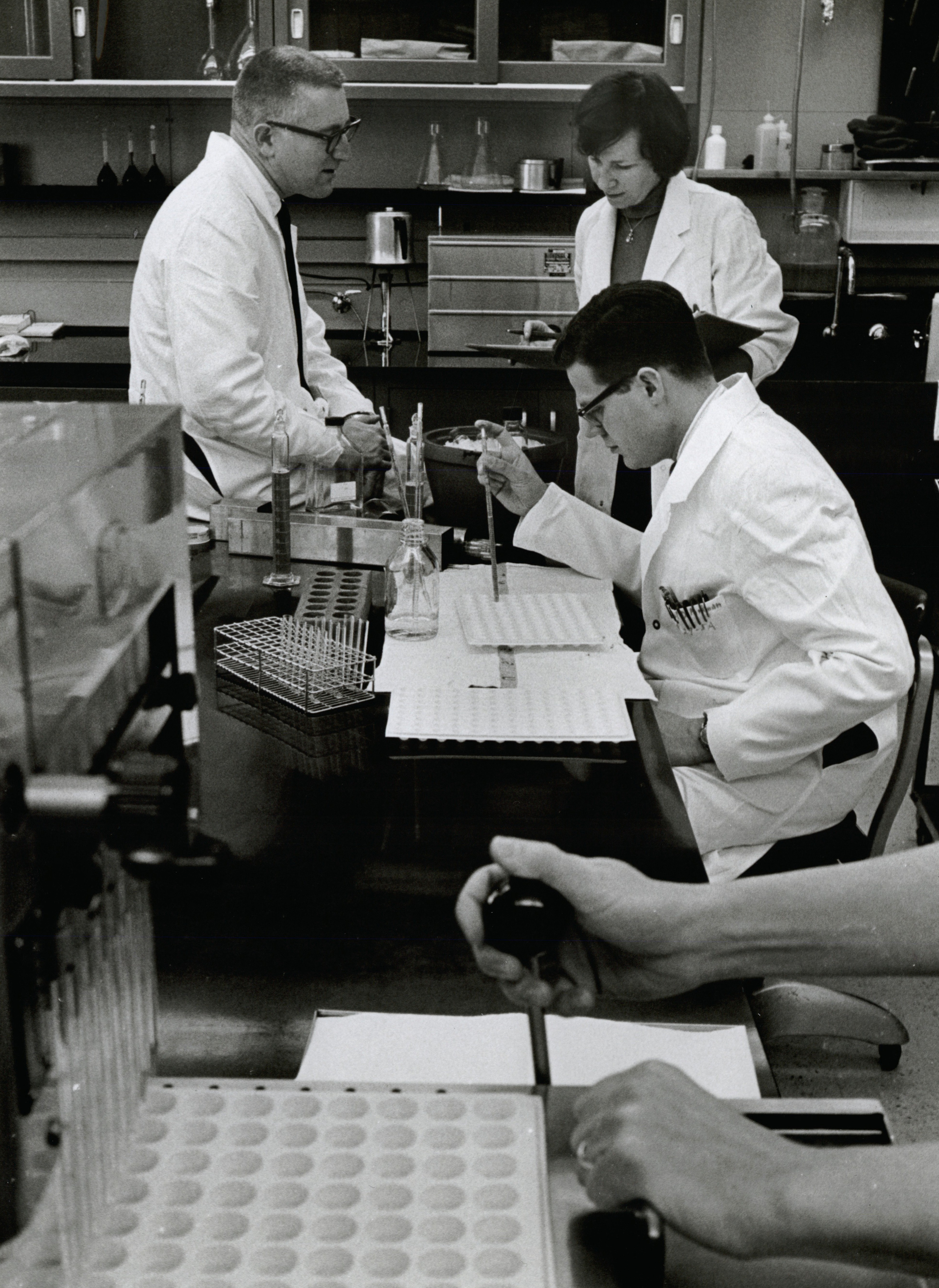a black and white photo of one woman and two men working in a lab, wearing white lab coats