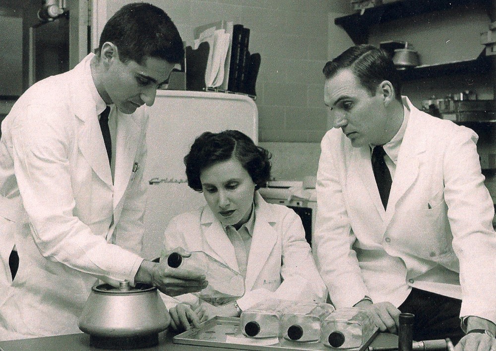 Three scientists in a laboratory looking at bottles of solution. The man on the left is holding up a bottle, a woman and a man are seated to the right. All are wearing white lab coats.
