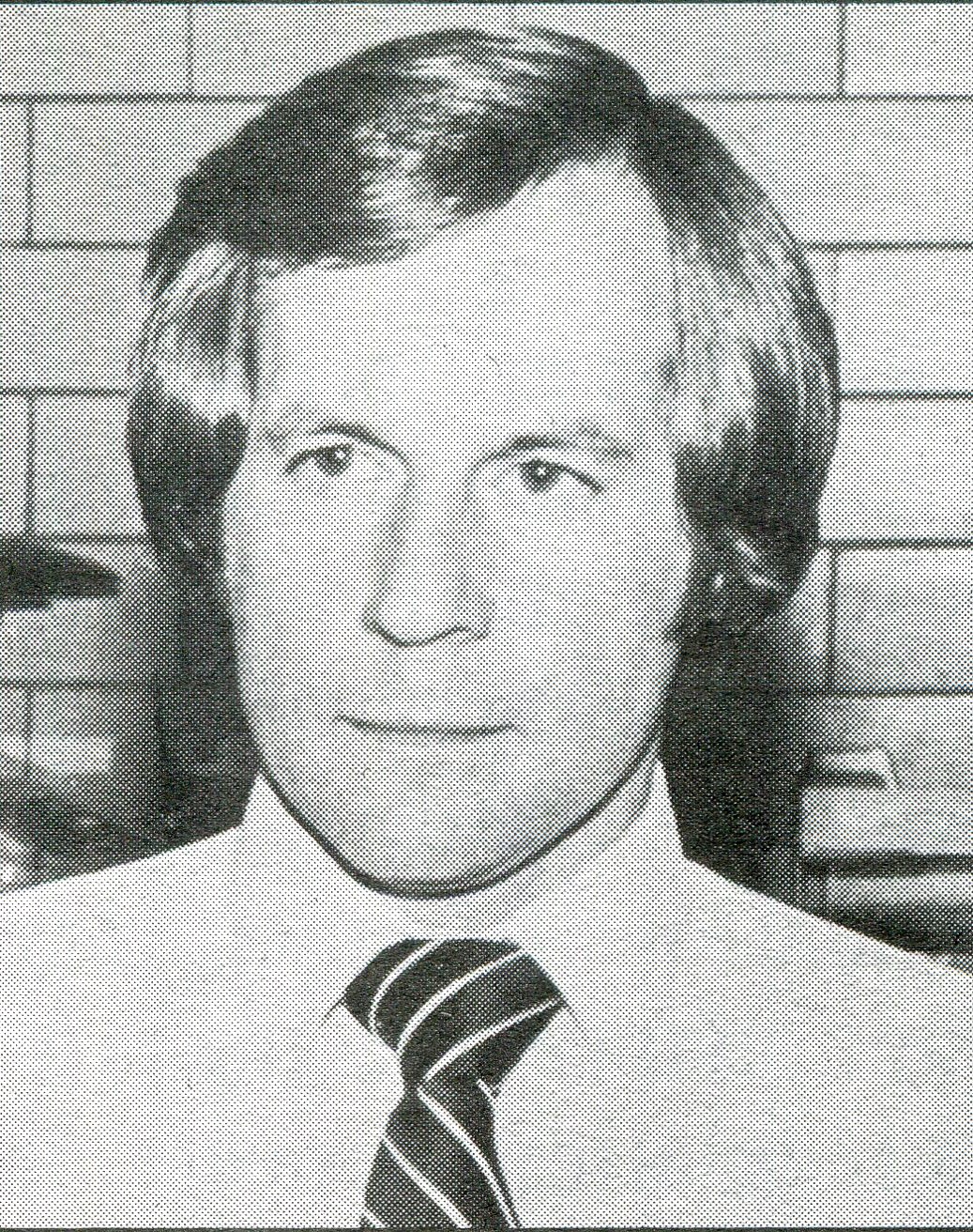 a black and white headshot of a man in a shirt and tie against a tile wall