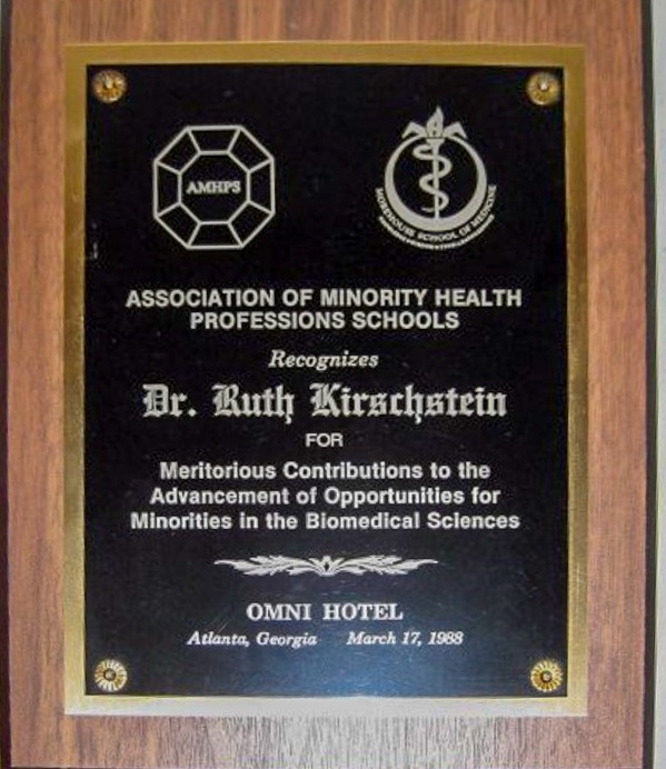 A photo of an award with a black plaque on faux wood grain. Presented to Ruth Kirschstein from the Association of Minority Health Professions Schools in 1988