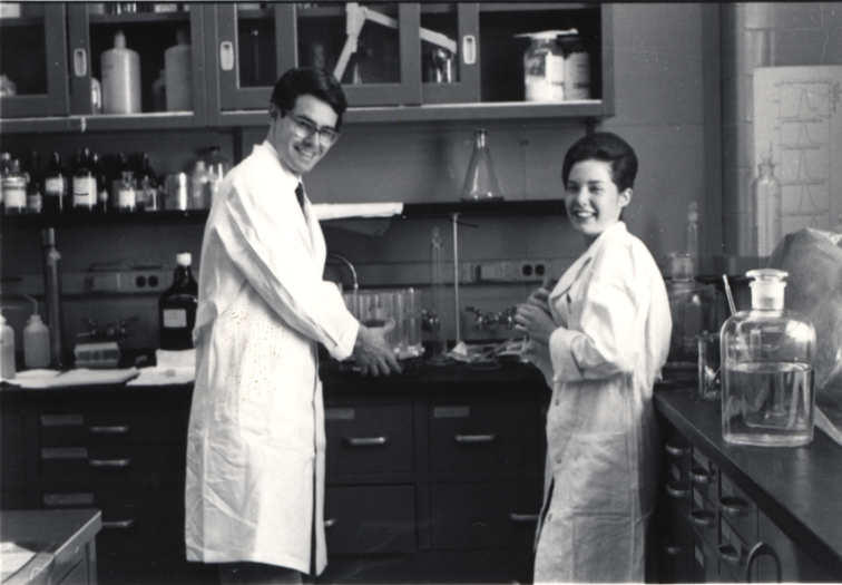 a man and woman scientist stand in a lab wearing white lab coats, looking at the camera smiling