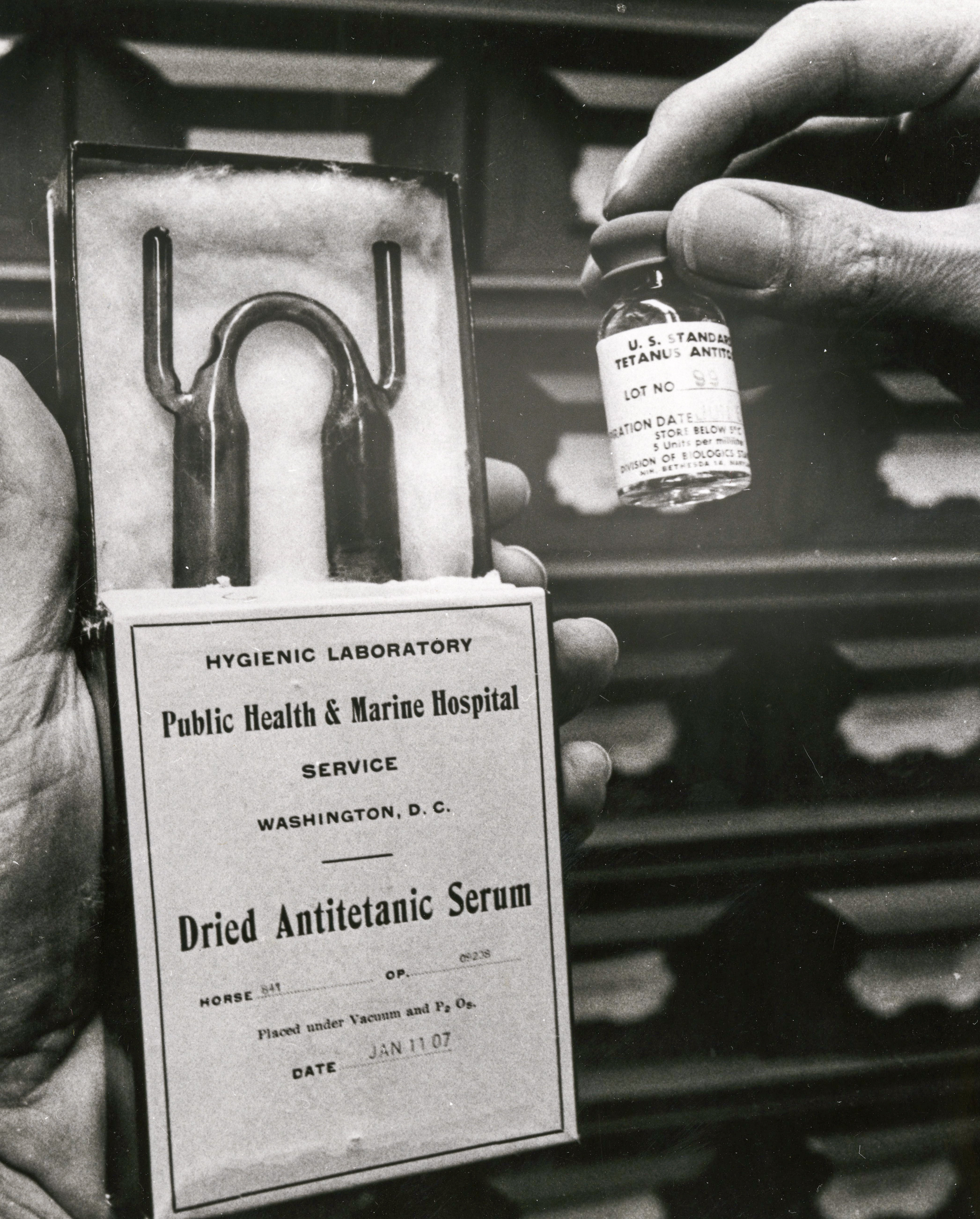 A vial of tetanus antitoxin and the dried antiserum are held up in someone's hands.