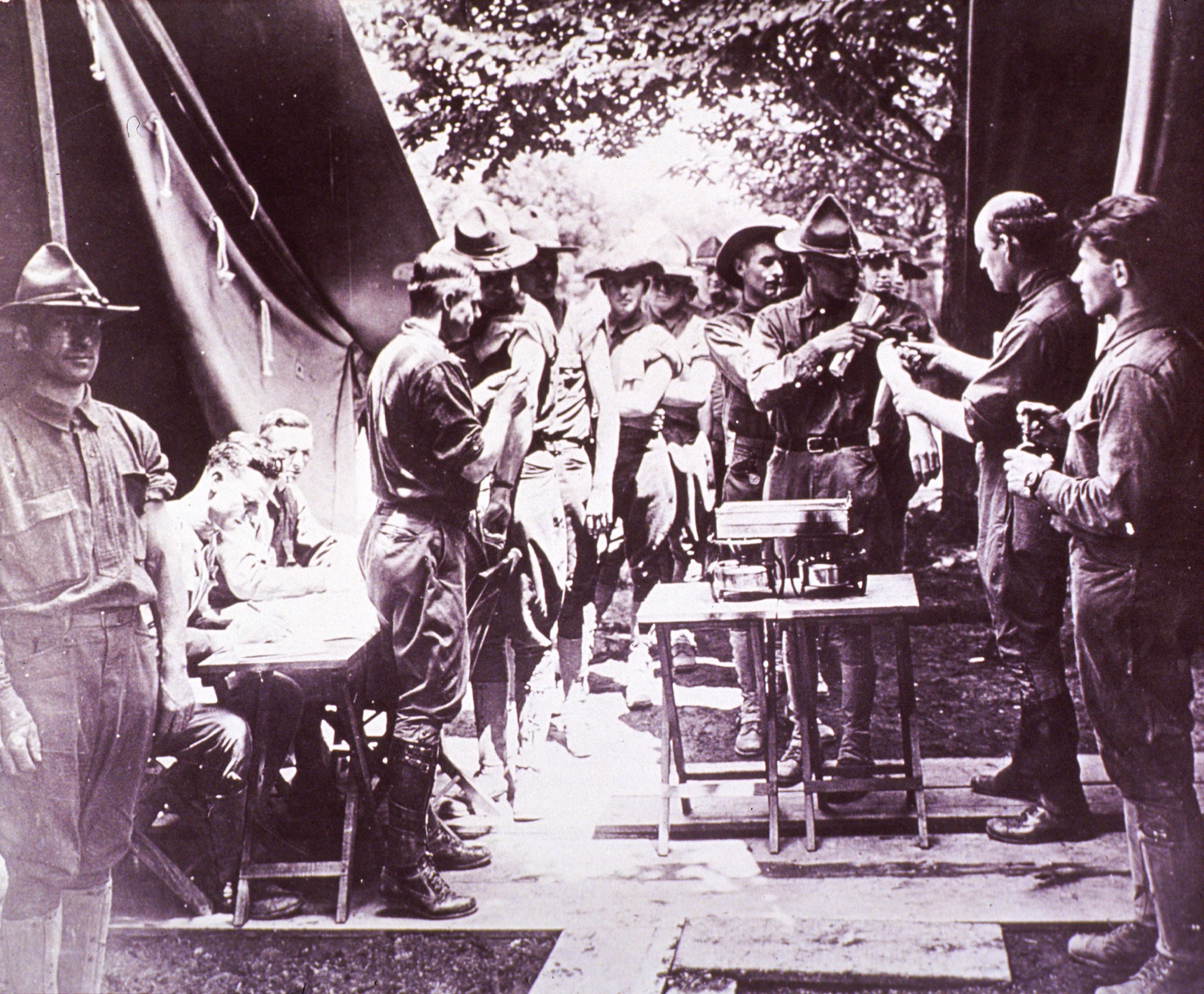 Black and white photograph of soldiers standing in two lines as the get vaccinated outdoors between two large tents and wood planks laid down as a walkway during World War I