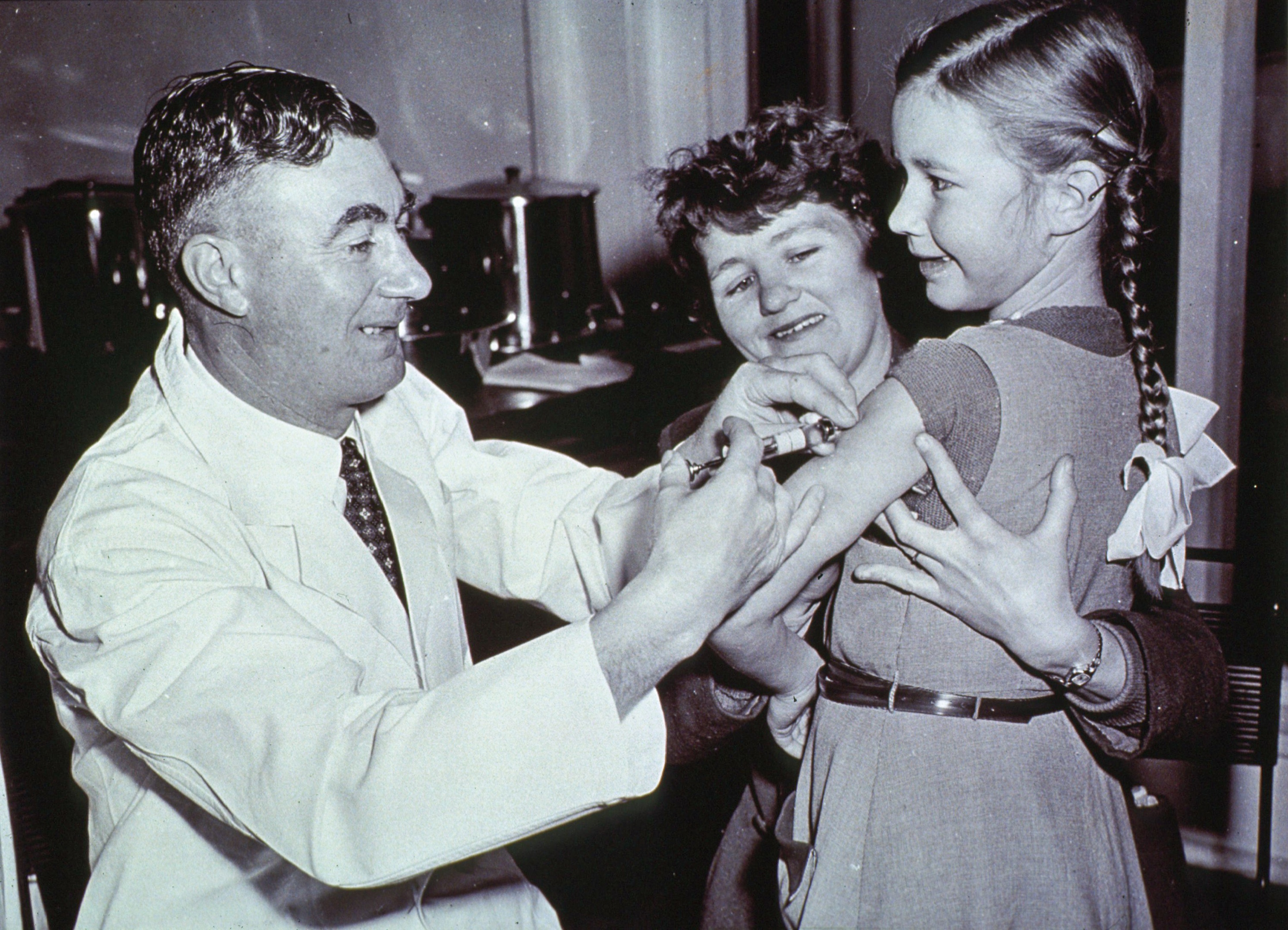 An Australian doctor is vaccinating a girl, who is smiling while a nurse looks on