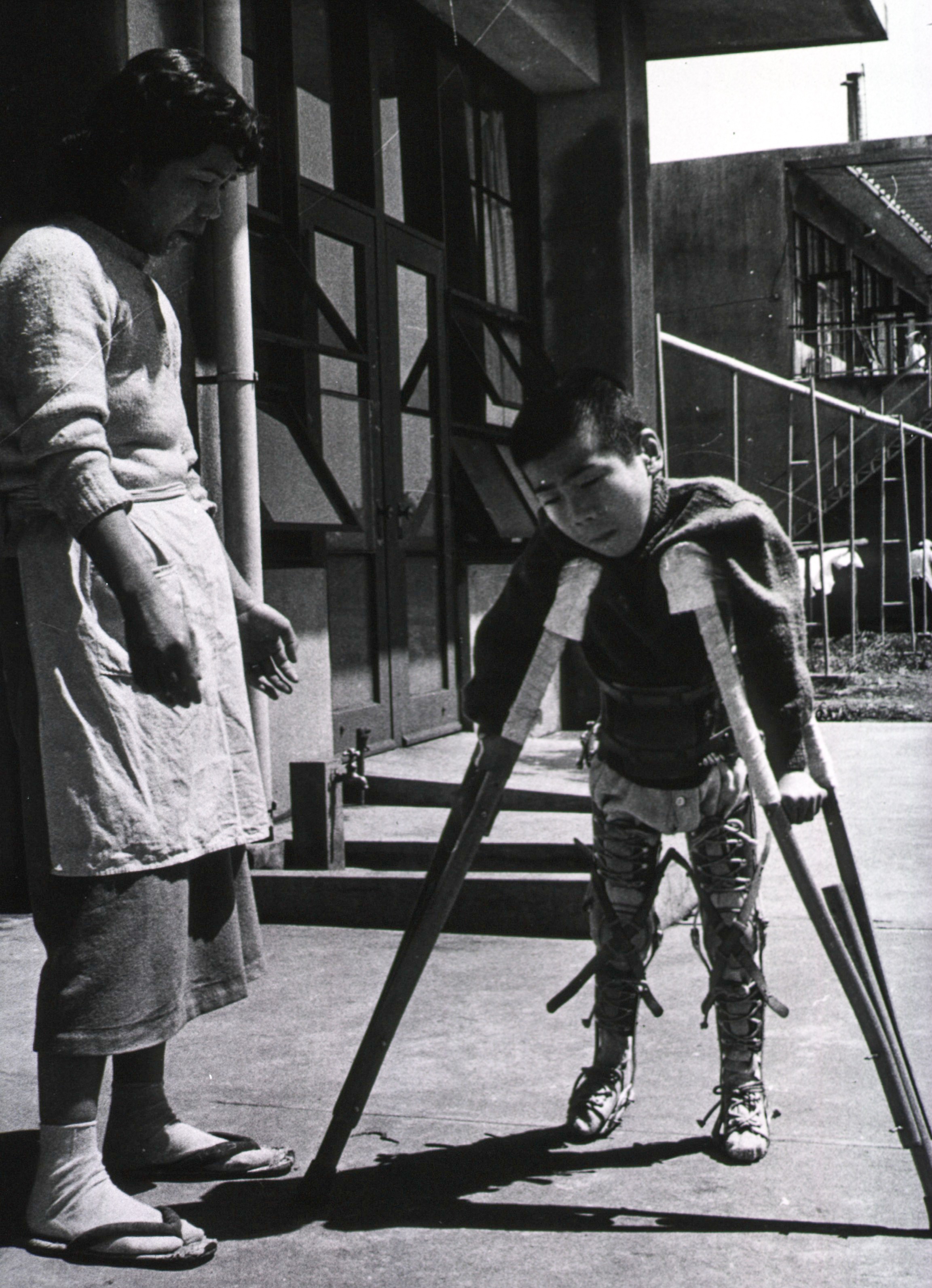 A black and white photo of a child on crutches trying to walk with a nurse nearby. Photo taken in Japan at a polio hospital