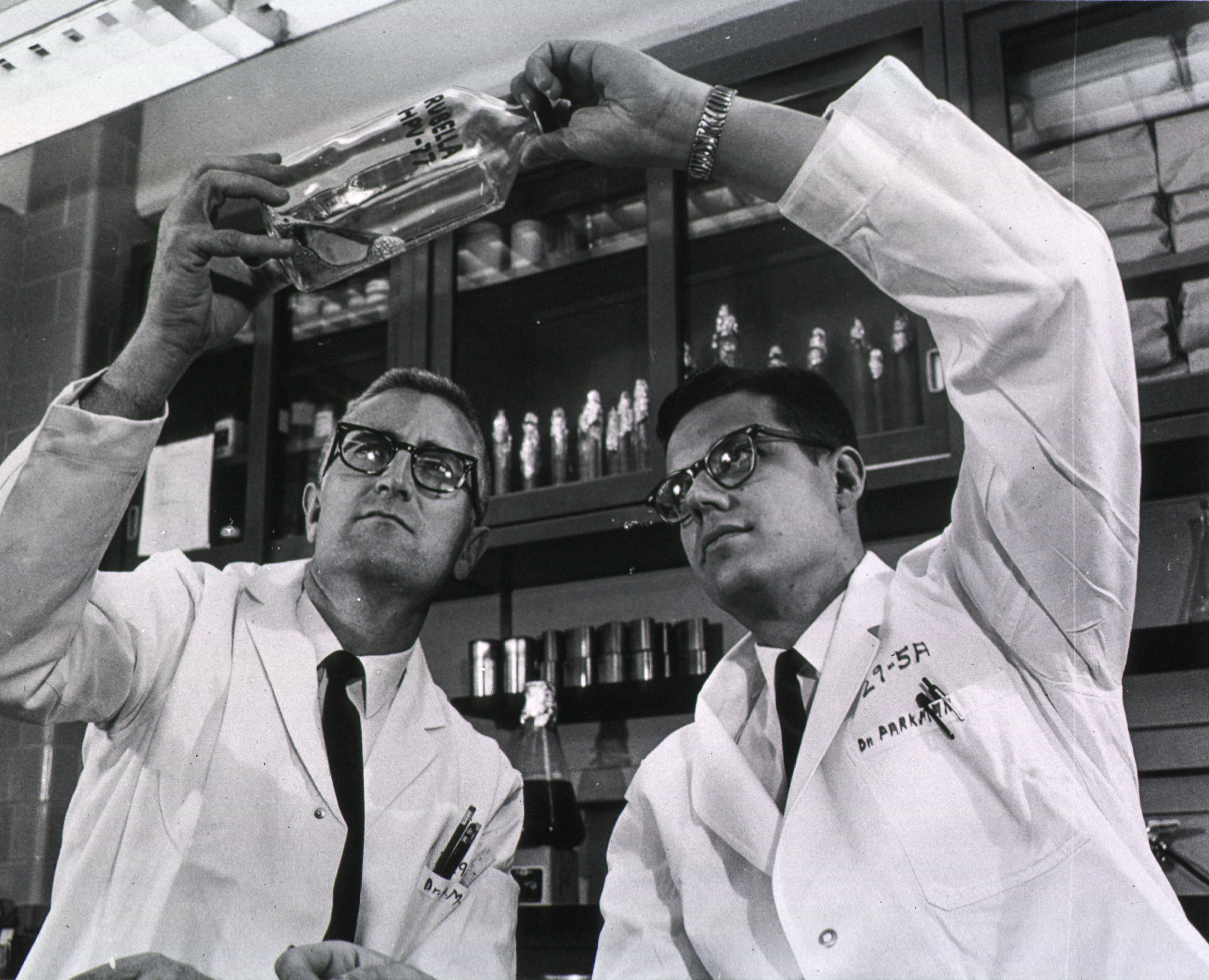Two men in lab coats examine a bottle labelled Rubella