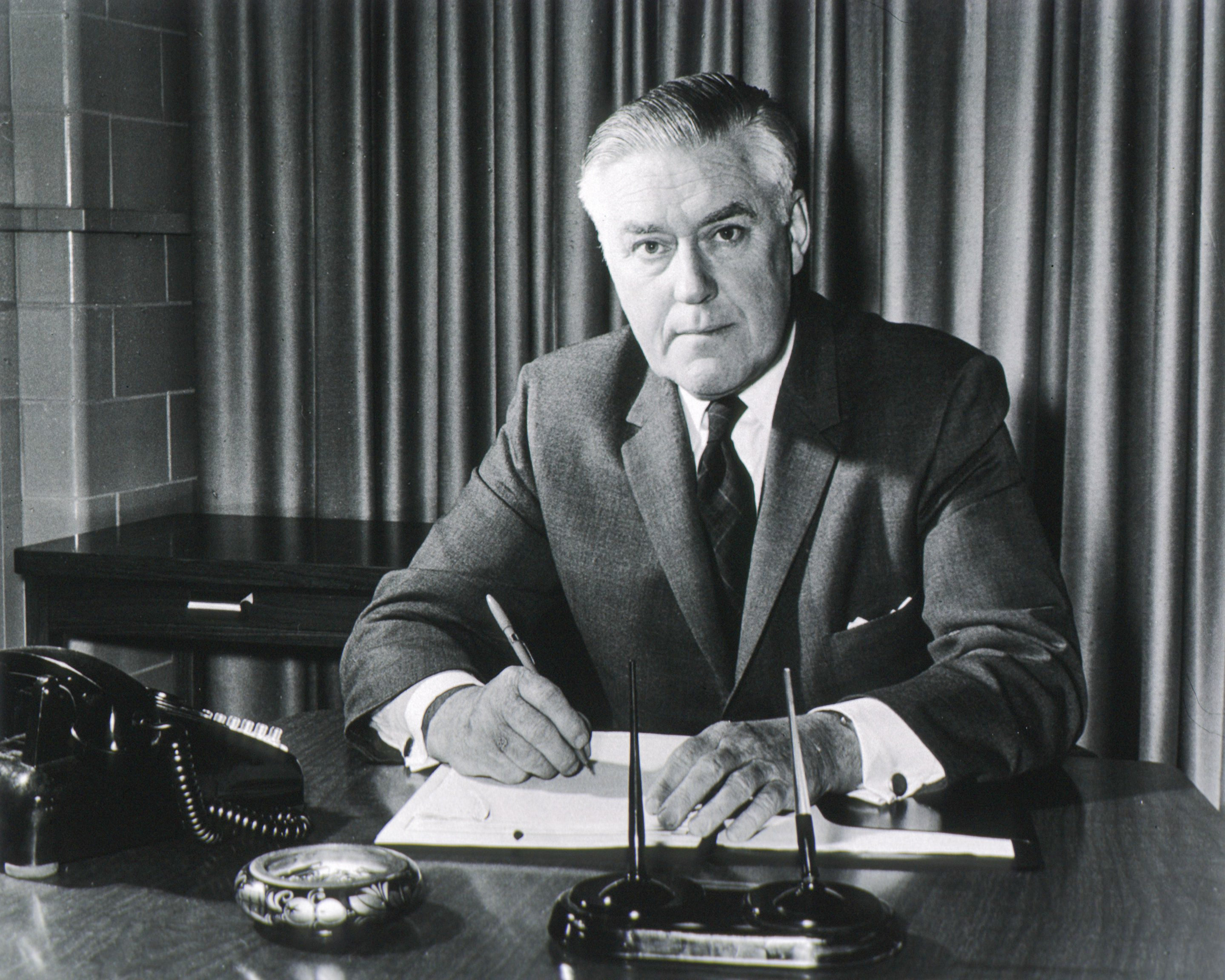 Dr Murray sitting at his desk wearing a suit, writing with a pen