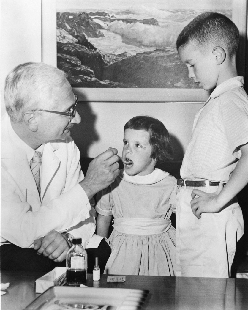 Dr. Sabin administers an oral vaccine to a girl in a dress next to an older boy