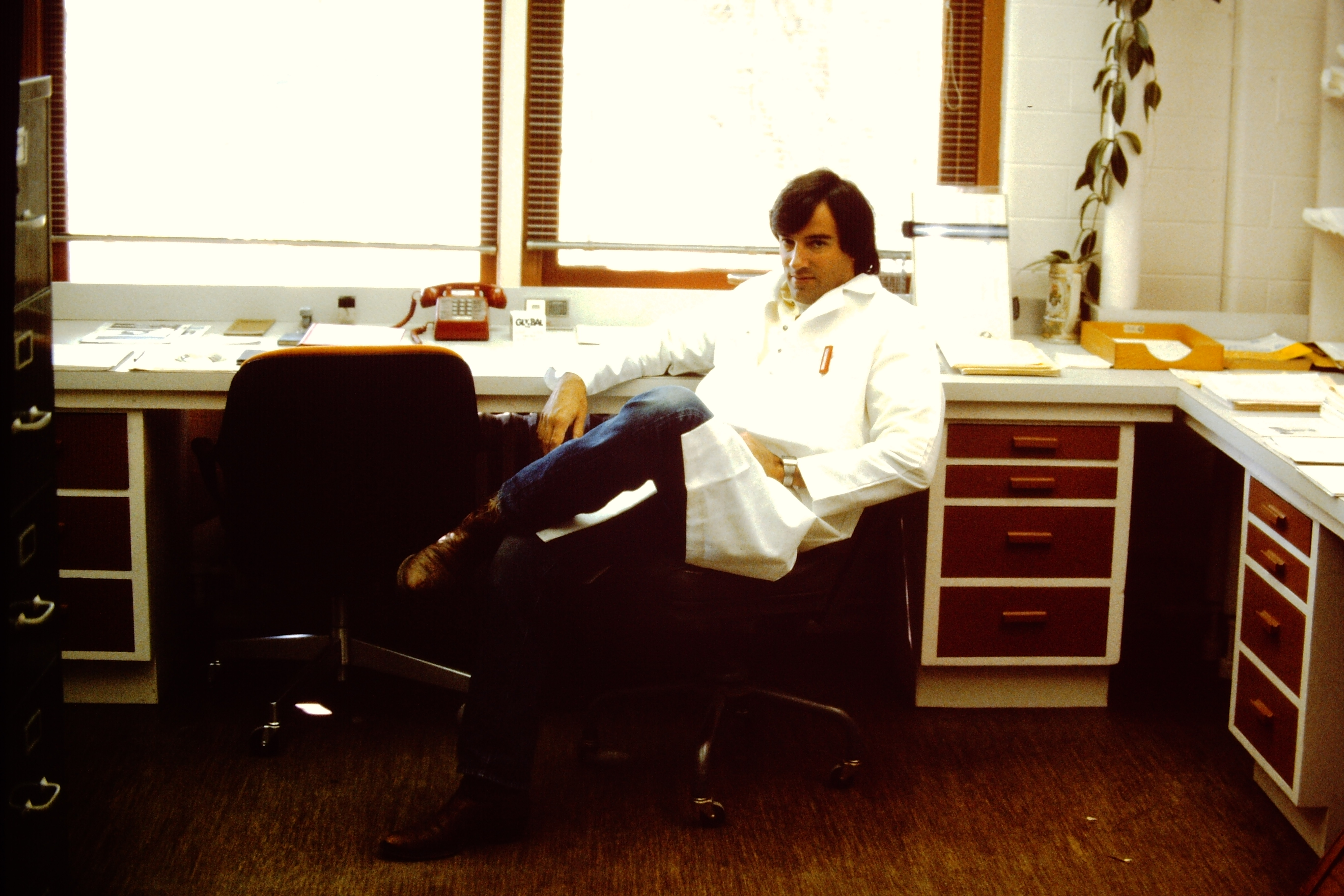 Man in white coat sits at desk in lab bench.