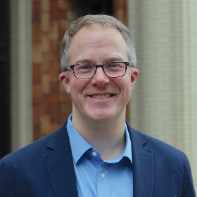 Dr. Christopher Phillips smiling man wearing glasses in light blue shirt and blue suit